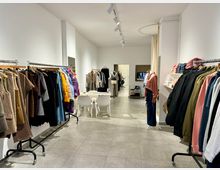 Ein moderner Kleidungsladen mit weißen Wänden und grauem Fliesenboden. Auf beiden Seiten des Raumes befinden sich Kleiderstangen, die mit farbenfroher Kleidung bestückt sind, sowie Regale mit gefalteten Kleidungsstücken; im Hintergrund sind ein Anprobe-Bereich und eine Tür zu sehen.