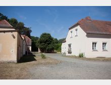 Hof mit historischen Gebäuden, gekennzeichnet durch cremefarbene Putzfassaden und rote Ziegeldächer. In der Mitte des Hofes befindet sich ein schmaler Kiesweg, umgeben von vereinzelten Bäumen und grasbewachsenen Flächen.