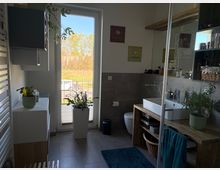 Modernes Badezimmer mit grauen Fliesen, großem Fenster bis zum Boden und Blick ins Freie. Rechts ein Waschtisch aus Holz mit offenem Regal, Wandregal mit Pflegeprodukten und Pflanzen sowie ein WC; vorne ist die Glaswand einer Dusche sichtbar.