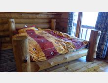 Holzinterior eines Schlafzimmers mit einem rustikalen Bett aus massiven Holzstämmen. Das Bett ist mit orange-roter Bettwäsche bezogen, daneben ist ein Fenster mit Vorhängen und ein Holzfußboden sichtbar.