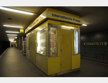 Gelber Kiosk mit der Aufschrift „Internationale Presse“ in einer U-Bahn-Station, umgeben von beleuchteten Schaufenstern mit Zeitschriften. Im Hintergrund ist der Stationsname „Westphalweg“ an einer dunklen gefliesten Wand zu sehen, mit Treppen und Hinweisschildern in Richtung Ausgang.