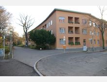 Wohngebäude mit orangefarbener Fassade und mehreren rechteckigen Balkonen, die teils mit Pflanzen dekoriert sind. Die Umgebung umfasst gepflasterte Gehwege, einige Bäume und eine ruhige Straßenkreuzung.