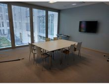 Ein moderner Besprechungsraum mit einem rechteckigen Tisch und sechs weißen Stühlen, einer grauen Wand mit einem angebrachten Flachbildschirm sowie einer großen Fensterfront mit Jalousien, die den Blick auf einen Innenhof ermöglicht. Der Boden ist mit Teppichboden ausgelegt, und an der Seite befindet sich ein kleiner Beistelltisch.
