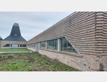 Modernes Gebäude mit einer markanten Ziegelfassade und großen rechteckigen Fenstern; im Hintergrund ein Turm mit einer schwarzen, spitz zulaufenden Dachkonstruktion. Die Umgebung besteht aus einer Grünfläche und einem unbebauten, teils bewachsenen Erdboden.