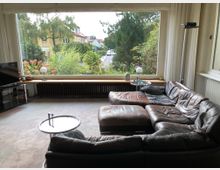 Wohnzimmer mit einer großen Panoramafensterfront, die auf eine grüne Straße mit Bäumen und gelben Häusern blickt. Innen befinden sich ein braunes Ledersofa in L-Form, ein kleiner runder Beistelltisch, ein Fernseher auf einem Glasregal sowie eine Fensterbank mit Dekorationen und Lampen.