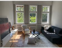 Helles Wohnzimmer mit großen Fenstern, die auf grüne Bäume blicken. Der Raum ist mit einem dunkelgrauen Sofa, einem Sessel mit Fellauflage, einem niedrigen Tisch, Kissen und modernen Möbeln eingerichtet.