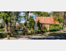 Ein kleines Backsteingebäude mit rotem Ziegeldach und teilweise von Efeu bewachsenen Wänden steht neben einer gepflasterten Straße, umgeben von Bäumen. Im Vordergrund ist ein schwarzes schmiedeeisernes Tor zwischen zwei gemauerten Pfeilern mit Kreuzen zu sehen, das den Eingang zu einem Friedhof markiert.