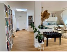 Heller Wohnbereich mit offener Gestaltung, bestehend aus einer Küche mit weißen Hochglanzschränken und schwarzen Arbeitsflächen, einem angrenzenden Essbereich mit Holztisch und weißen Stühlen. Links steht ein Regal voller Bücher, in der Mitte ein Tischkicker, und Pflanzen sowie Dekorationselemente verleihen dem Raum einen wohnlichen Charakter.