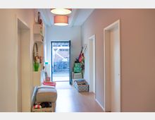 Moderner Flur mit Holzboden, weißen Wänden und einer offenen Tür zum Garten. Links befinden sich Wandregale mit Dekorationen und Stauraum, rechts eine Garderobe mit Haken und Körben für diverse Gegenstände.