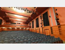 Großer Innenraum eines Theaters oder Konzerthauses mit mehreren Sitzreihen, einer oberen Galerie und Holzverkleidungen an den Wänden. Über der Bühne befinden sich Scheinwerfer und Lautsprecher, die für die Beleuchtung und Beschallung installiert sind.