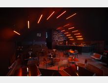 Dunkel beleuchtete Bar mit modernem Design, in der Decke in Rot- und Orangetönen leuchtende Lampen angebracht sind. In der Mitte befinden sich kleine Tische mit Kerzen, im Hintergrund eine Theke mit Barhockern und einer Regalwand für Getränke.