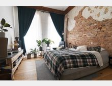 Schlafzimmer mit einem Bett im Vordergrund, bedeckt mit karierten Decken, und einer Ziegelsteinwand im Hintergrund. Große Fenster mit dunklen Vorhängen lassen Tageslicht herein; Pflanzen stehen auf Regalen und Tischen, und ein Sessel steht in der Ecke.