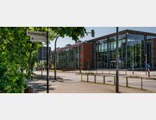 Moderne Straßenansicht mit einem großen Gebäude aus Glas und rotem Backstein auf der rechten Seite, umgeben von Gehwegen und Straßenlaternen. Im Vordergrund befindet sich ein Wegweiser mit der Aufschrift „Mühlenstraße“ sowie grüne Bäume, die Schatten spenden.