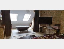 Dachgeschosszimmer mit freistehender schwarzer Badewanne auf einem weißen Podest, zwei Dachfenstern, einer unverputzten Backsteinwand und einem großen Fernseher auf einem Holztisch. Im Hintergrund steht ein dekoratives Kleidermodell neben einem dunklen Vorhang.