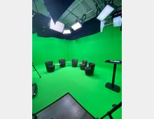 Ein Studio mit grünem Hintergrund (Green Screen) und mehreren Studiolampen an der Decke. Im Raum stehen fünf schwarze Sessel und drei kleine Beistelltische in einem Halbkreis, daneben ein Stehpult.