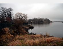Flussuferlandschaft an einem trüb bewölkten Tag mit kahlen Bäumen, einem kleinen Bootsanleger und trockenem, gelblich-braunem Gras im Vordergrund. Der Fluss erstreckt sich ruhig, gesäumt von Bäumen und Sträuchern.