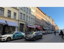 Straßenszene in einer Stadt mit Reihen historischer Wohn- und Geschäftshäuser; im Erdgeschoss befinden sich kleine Geschäfte mit Markisen, davor parkende und fahrende Autos sowie Fahrradfahrer. Der Himmel ist klar und blau.