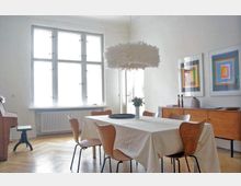 Speisezimmer mit großem Tisch und Holzstühlen, dekoriert mit einer weißen Tischdecke und einer Vase mit Blumen. Im Hintergrund ein großes Fenster, ein hölzernes Sideboard mit Kunstbildern an der Wand sowie ein auffälliger, weißer Hängelampen-Schirm.