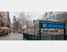 Straßenszene am Kurfürstendamm in Berlin mit einem U-Bahneingang der Linien U1 und U9 im Vordergrund, umgeben von Geschäften und moderner Architektur. Im Hintergrund ist die Kaiser-Wilhelm-Gedächtniskirche und eine mit kahlen Bäumen gesäumte Straße sichtbar.