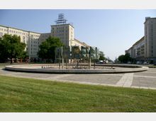 Ein runder, leerer Brunnen mit einem modernen Kunstwerk in der Mitte, bestehend aus metallischen, geometrischen Elementen, umgeben von einer gepflasterten Fläche und Rasen. Im Hintergrund stehen symmetrische, mehrstöckige Gebäude im klassischen sozialistischen Baustil, gesäumt von Bäumen und einer Straße mit Autos.