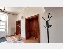 Flur mit hellen Wänden und einer weißen Eingangstür mit Glaseinsätzen links. In der Mitte befinden sich zwei Holzrahmentüren, ein Heizkörper und ein Feuerlöscher. Zwei Garderobenständer stehen auf einem gefliesten Boden in beige-weißem Muster.