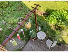 Kleiner Garten mit einer Holzterrasse, auf der ein runder Metalltisch und zwei Metallstühle stehen. Darüber befindet sich ein Holzgerüst mit bunten Dekorationen und einer Papierlaterne, umgeben von dichter Vegetation und Blumen.