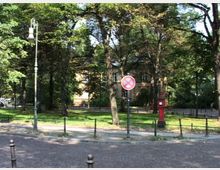 Parkähnliche Umgebung mit gepflasterter Straße im Vordergrund, Bäumen und Grünflächen. Im Hintergrund steht ein helles Gebäude, flankiert von einem historischen Laternenmast, einem Verkehrsschild und einer roten Säule.