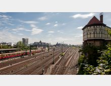 Blick auf ein großes Bahnhofsareal mit zahlreichen Gleisen und Oberleitungen, links ein Bahnsteig mit abgestellten S-Bahn-Wagen. Rechts steht ein historisches Gebäude mit Fachwerk und rotem Dach, im Hintergrund sind städtische Gebäude und Industrieanlagen zu sehen.
