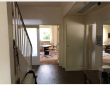 Innenaufnahme eines Flurs mit Treppe auf der linken Seite, der zu einem hellen Raum mit großen Fenstern und einem Tisch mit Stühlen führt. Rechts sind weitere Türen und ein Wandspiegel sichtbar.