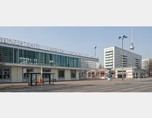 Straßenansicht des "Café Moskau" in Berlin, einem flachen Gebäude mit großen Glasfassaden und einem Schriftzug auf dem Dach. Im Hintergrund sind Wohngebäude und der Berliner Fernsehturm sichtbar, während im Vordergrund eine Bushaltestelle zu sehen ist.