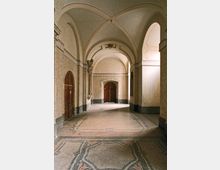 Ein historischer Flur mit hohen Gewölbedecken, verziertem Stuck und hellen Wänden. Der Boden ist mit einem kunstvollen Muster aus Steinfliesen gestaltet, Türen aus dunklem Holz säumen die Seiten, und am Ende des Flurs befindet sich eine doppelflügelige Holztür.