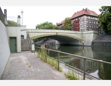 Fußweg entlang eines Kanals mit einer Betonbrücke im Hintergrund. Links führt eine Treppe zu einer höheren Ebene mit einer Tür, und rechts sind Geländer sowie vereinzelte Pflanzen sichtbar. Im Hintergrund befinden sich mehrstöckige Gebäude mit roten Dächern und grüner Vegetation.