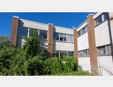 Mehrstöckiges Büro- oder Wohngebäude mit einer Fassade aus weißen und braunen Elementen, großen rechteckigen Fenstern und wuchernder Vegetation im Vordergrund. Der Himmel ist klar und strahlend blau.