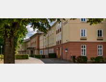 Historisches Gebäude mit hellgelber und terrakottafarbener Fassade, großen Fenstern und dekorativen Details. Der Eingangsbereich hat ein breites Vordach, im Vordergrund ist ein gepflasterter Weg mit Bäumen und Sträuchern sichtbar.