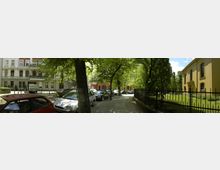 Straßenszene mit parkenden Autos unter grünen Bäumen. Rechts ein gelbes Gebäude mit hohen Fenstern hinter einem schwarzen Metallzaun, links Wohnhäuser mit dekorativen Fassaden.