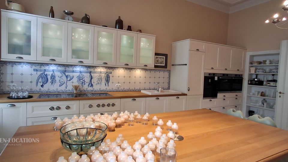 Große, helle Wohnküche mit weißen Einbauschränken und Milchglas-Oberschränken, Holzarbeitsplatte und blau-weiß gefliestem Fliesenspiegel mit dekorativen Motiven. Im Vordergrund steht eine große Kücheninsel aus Holz; rechts sind Hochschränke mit Einbauöfen und ein offenes Regal neben einer weißen Tür.