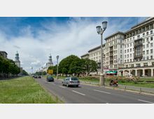 Straßenszene in der Karl-Marx-Allee in Berlin, gesäumt von großen, symmetrischen Gebäuden im Stil des sozialistischen Klassizismus. Im Hintergrund sind die Türme der Frankfurter Tor sowie der Fernsehturm sichtbar, während Bäume, Straßenlaternen und Fahrzeuge die Allee prägen.