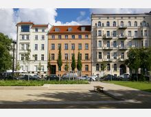 Straßenszene mit historischen Wohngebäuden im Vordergrund; links ein weißes Gebäude, in der Mitte ein orangefarbenes, rechts ein beiges Gebäude mit Sichtbalkonen, davor parkende Autos und ein kleiner Park mit Bäumen, Grünflächen und Sitzbänken.