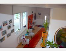 Wohnbereich mit heller Einrichtung, bestehend aus zwei Fenstern mit halbtransparenten Rollos, dekorativen Bildern an der Wand, einer kleinen Sitzgruppe mit Tisch, grünen Pflanzen und einem Teppichläufer. Im Hintergrund führen eine weiße Eingangstür und rote Holztüren zu anderen Räumen.