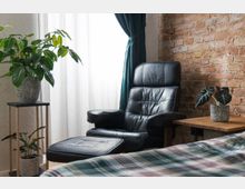 Schlichte Wohnzimmerecke mit einem schwarzen Ledersessel, passender Fußstütze und Holztisch, auf dem eine Topfpflanze steht; links ein Beistelltisch mit weiteren Pflanzen, vor einem weißen Vorhang und einer freiliegenden Ziegelwand.