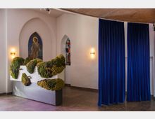 Ein Innenraum mit weiß verputzten Wänden, gotischen Spitzbögen und einem Mosaikfenster mit einer Heiligendarstellung. In der Mitte steht eine moderne Skulptur mit Moos und Pflanzen, daneben hängen dunkelblaue Vorhänge.