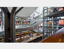 Innenansicht eines modernen Bahnhofs mit mehreren Ebenen, Rolltreppen und verglasten Aufzügen. Unten befinden sich Gleise und Bahnsteige, oben Einkaufsbereiche und Beschilderungen.