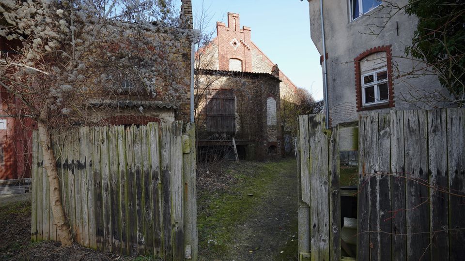 Blick durch ein geöffnetes, verwittertes Holztor in einen schmalen Hofweg zwischen alten Gebäuden aus Backstein und Putz mit roten Ziegelumrahmungen. Im Hintergrund stehen eine ziegelgedeckte Scheune und weitere Backsteinbauten, umgeben von kahlen Sträuchern und Bäumen.
