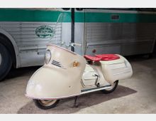 Beiger Motorroller im Retrodesign mit hoher, transparenter Windschutzscheibe und rotem Sitz, abgestellt auf Betonboden. Im Hintergrund steht ein silber-grüner Bus mit genieteter Metallverkleidung.