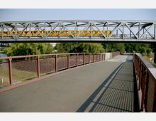 Eine metallene Fußgängerbrücke mit braunen Geländern überspannt einen Fluss, im Hintergrund verläuft eine Eisenbahnbrücke mit einem gelben Zug. Im Umfeld befinden sich grüne Bäume und ein gepflasterter Weg.