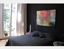 Schlafzimmer mit dunkler Wand, auf der ein abstraktes Gemälde hängt. Ein Bett mit dunkler Bettwäsche steht vor der Wand, daneben ein kleiner Nachttisch, und eine Glastür mit bodenlangen Vorhängen führt zu einer Terrasse mit Pflanzen.