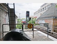 Bahnhofsumgebung mit Blick auf Gleise, eine einfahrende S-Bahn und ein Signal mit rotem Licht. Links und rechts befinden sich Graffiti-bedeckte Gebäude und eine Leitplanke mit einem Warnschild, das das Betreten der Gleise untersagt.