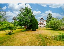 Großer Garten mit Rasenfläche und mehreren Bäumen, im Hintergrund ein weißes Fachwerkhaus mit spitzem Giebel und roten Dachziegeln. Der Himmel ist wolkig, und das Gelände wirkt gepflegt und ländlich.