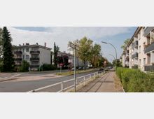 Straßenansicht eines Wohngebiets mit mehrstöckigen Wohnhäusern, bepflanzten Balkonen und Gehwegen, die von einem niedrigen weißen Geländer abgegrenzt sind. Im Hintergrund sind Bäume, parkende Autos und Straßenschilder zu sehen.