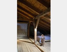 Dachboden aus Holz mit sichtbaren Balken, Holzboden und geneigtem Dach. Im Raum befinden sich ein Bett, ein Spinnrad sowie ein Vorhang mit dekorativem Muster, der eine Ecke abschirmt.